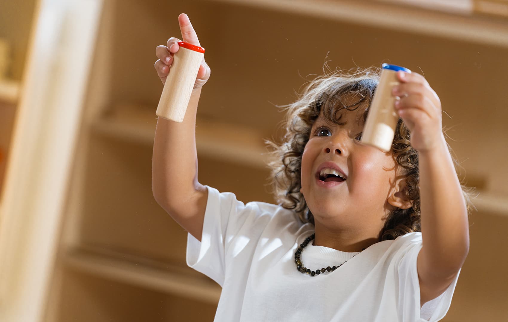 An enthusiastic child holding two wooden Nienhuis Sound box tubes, each with a colored top, in a Montessori environment.