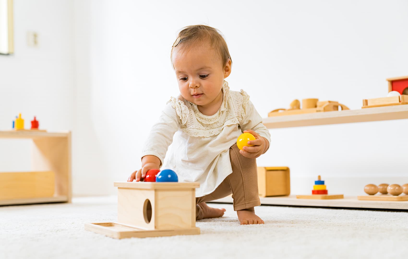 A young child kneeling with the Montessori product 'Push Box' and concentrating on placing balls into a wooden box with round holes in a bright Montessori space.